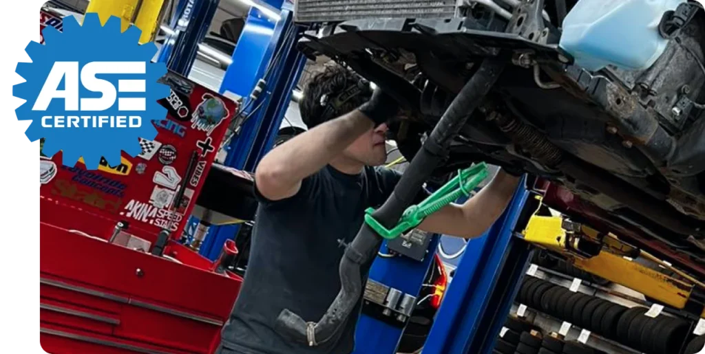 A mechanic working under a car in a garage with an "ASE Certified" logo in the top left corner.