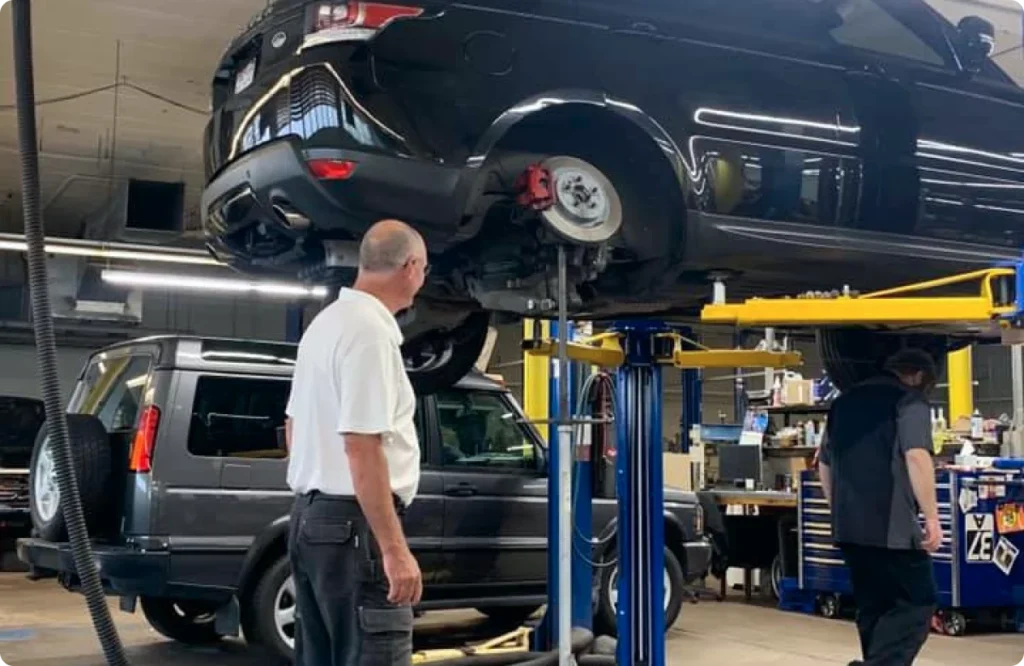 Mechanics work in an auto repair shop with a black SUV lifted on a hydraulic lift for servicing.