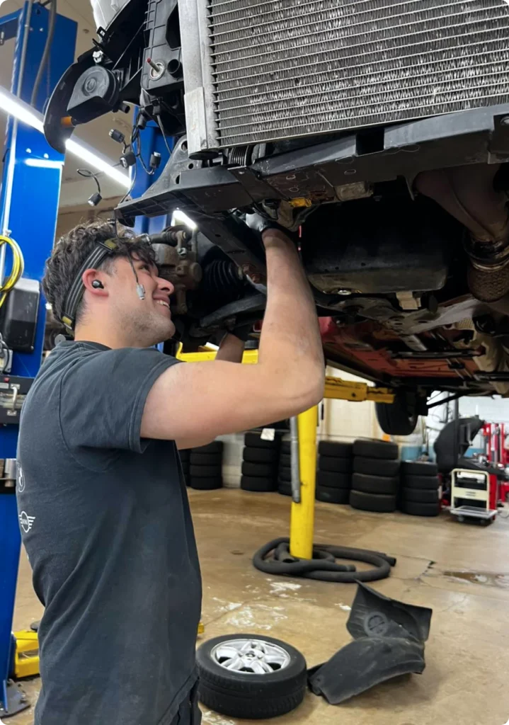 Mechanic smiling while working underneath a lifted car in an automotive repair shop.