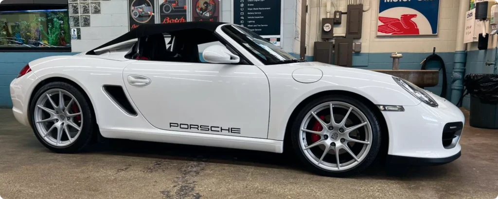 A white Porsche convertible parked in a garage in Milwaukee, WI, with automotive posters and tools in the background.