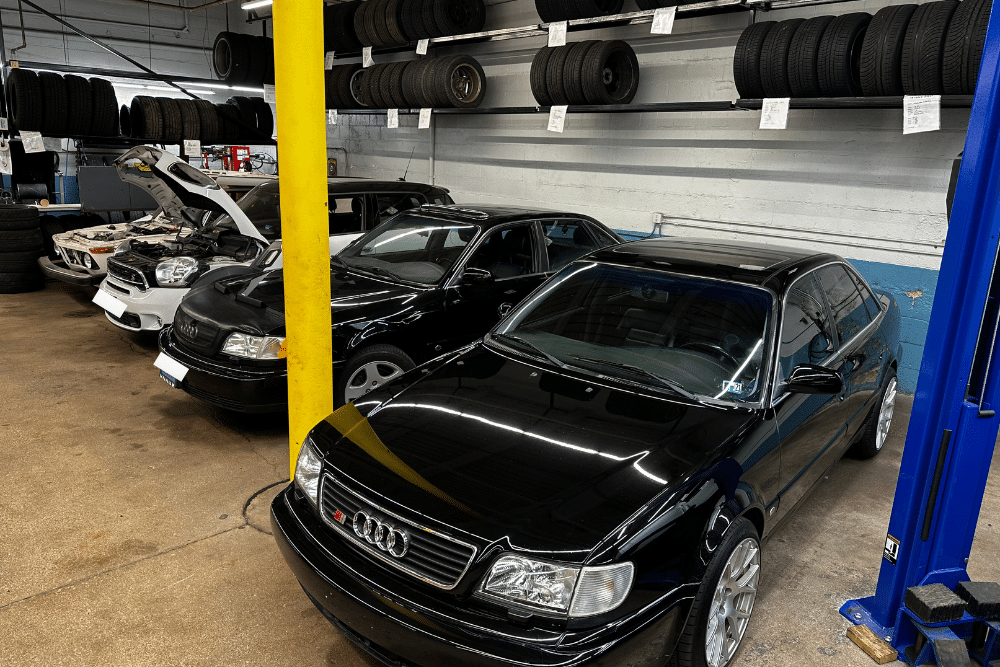 audi engine repair, european auto repair in Milwaukee, WI at O'Reilly Motor Cars. Automotive repair shop interior with several cars parked inside, one with its hood open, and tires stacked on racks above.