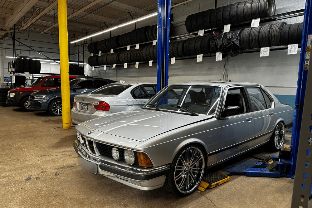 BMW Leak Oil, european auto repair in Milwaukee, WI at O'Reilly Motor Cars. Classic silver BMW on a lift in an auto shop with other cars and tires in the background.