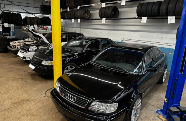 audi engine repair, european auto repair in Milwaukee, WI at O'Reilly Motor Cars. Automotive repair shop interior with several cars parked inside, one with its hood open, and tires stacked on racks above.