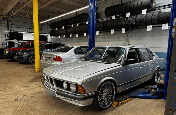 BMW Leak Oil, european auto repair in Milwaukee, WI at O'Reilly Motor Cars. Classic silver BMW on a lift in an auto shop with other cars and tires in the background.